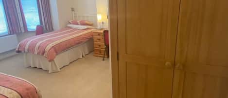 A bedroom with two beds, a wooden wardrobe, a bedside table with a lamp, and a window with curtains.