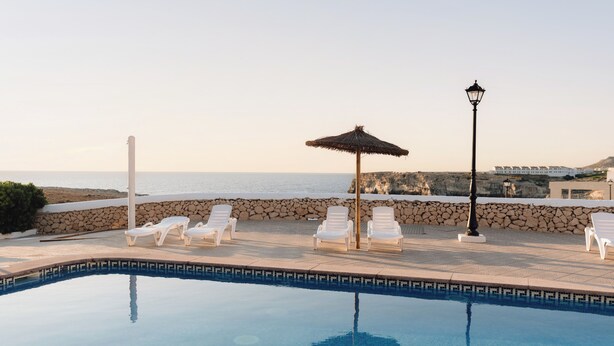 A pool area with lounge chairs and a thatched umbrella, overlooking a coastal landscape. - Bluesea Cabo de Baños (Ciutadella de Menorca)