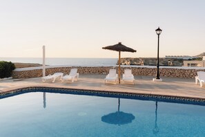 A pool area with lounge chairs and a thatched umbrella, overlooking a coastal landscape. - Bluesea Cabo de Baños (Ciutadella de Menorca)