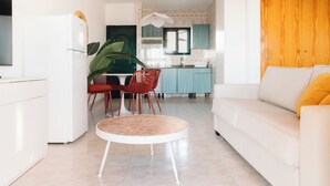 A modern living room with a white sofa and a round coffee table, a kitchen with teal cabinets and a window, and a dining area with red chairs and a plant. - Bluesea Cabo de Baños (Ciutadella de Menorca)