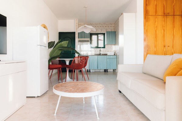 A modern living room with a white sofa and a round coffee table, a kitchen with teal cabinets and a window, and a dining area with red chairs and a plant. - Bluesea Cabo de Baños (Ciutadella de Menorca)