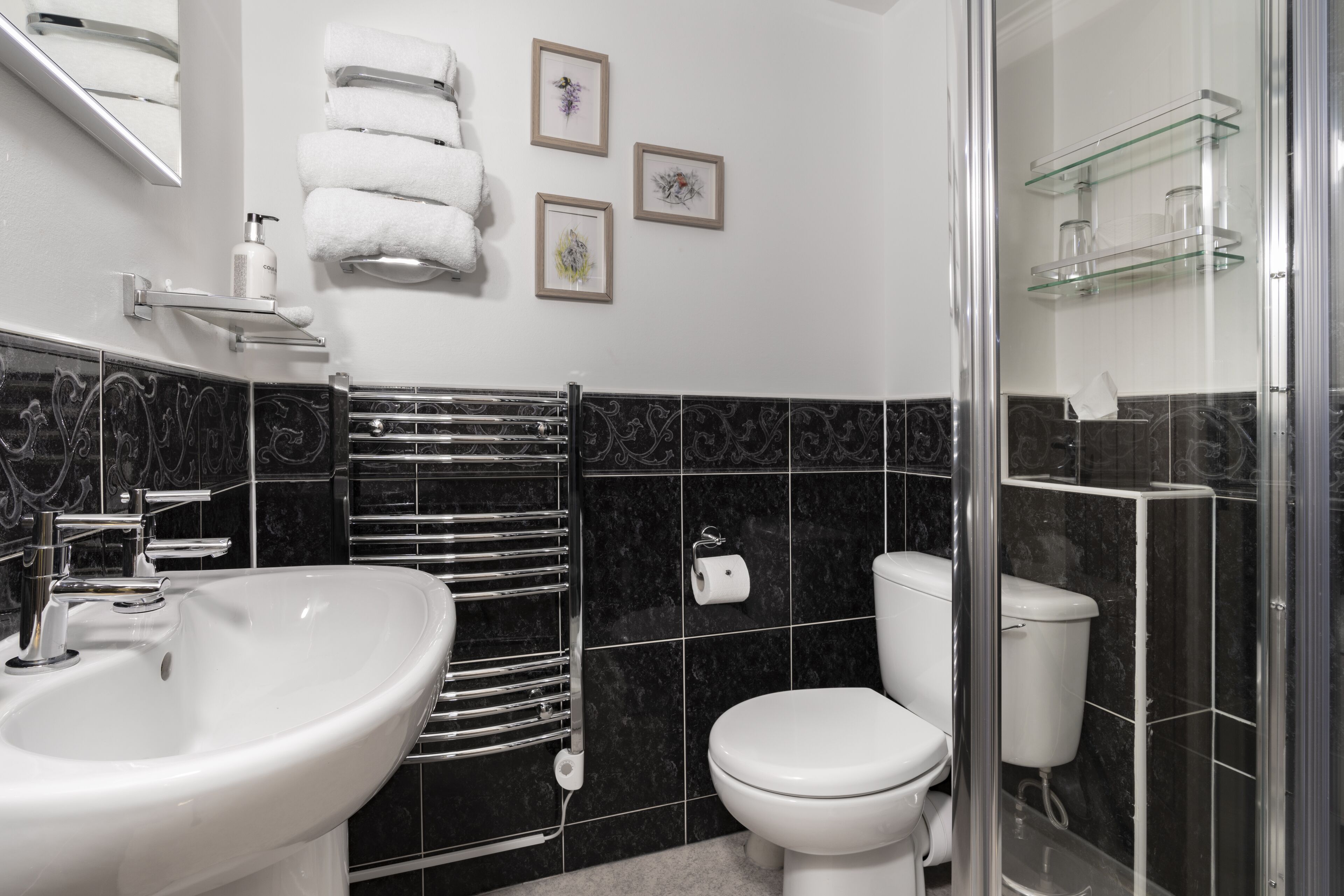 A modern bathroom with a white sink, chrome towel rack, black marble tiles, a toilet, and framed pictures on the wall.