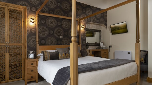 A four-poster bed with a wooden frame, a nightstand with drawers, a bedside table, a mirror, and a television on the wall. - Melrose Guest House (Ambleside)