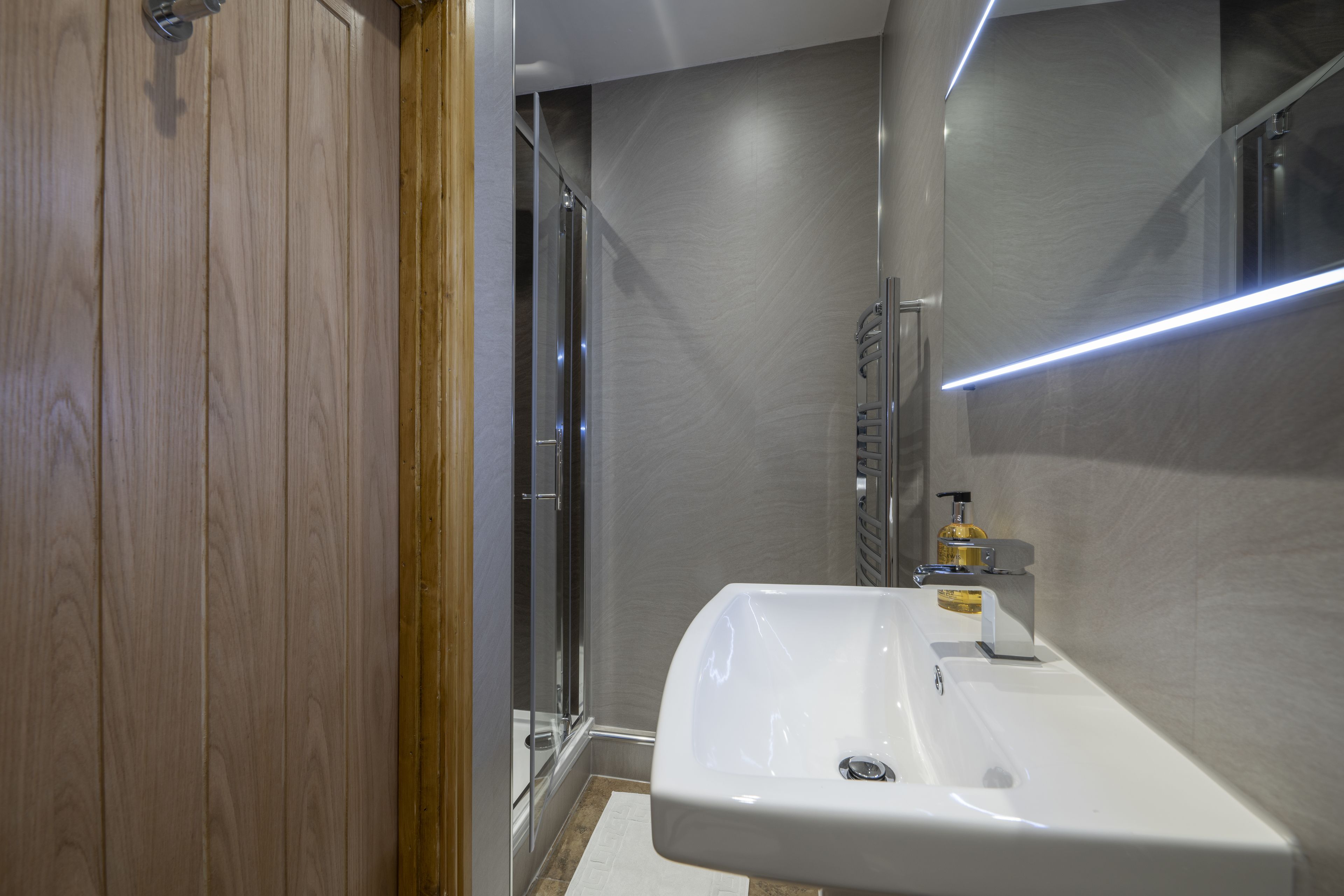 A modern bathroom with a wooden door, a glass shower enclosure, and a white sink with a soap dispenser.