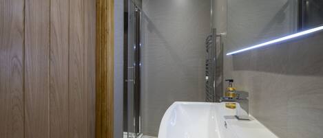 A modern bathroom with a wooden door, a glass shower enclosure, and a white sink with a soap dispenser.