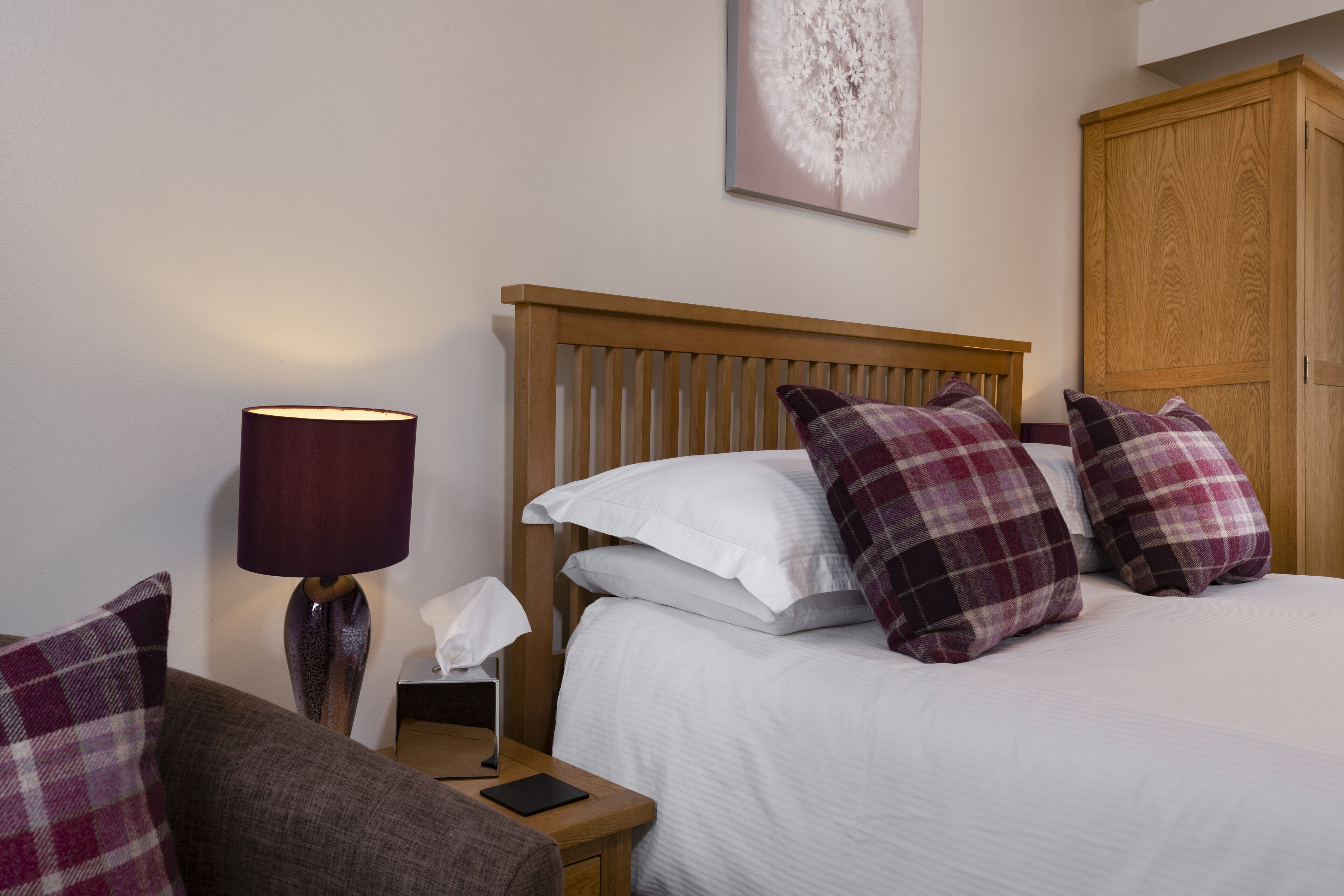 Een net opgemaakt bed met witte en geruite kussens, een houten hoofdbord, een bedlampje en een klein nachtkastje met een zakdoosje en een boek.