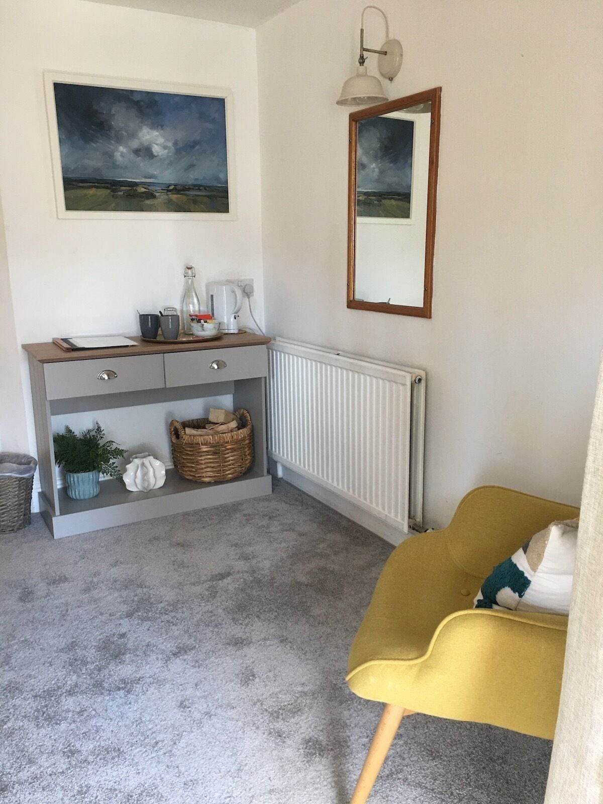 Une pièce avec une console grise, une chaise jaune, un radiateur et deux tableaux encadrés accrochés au mur.
