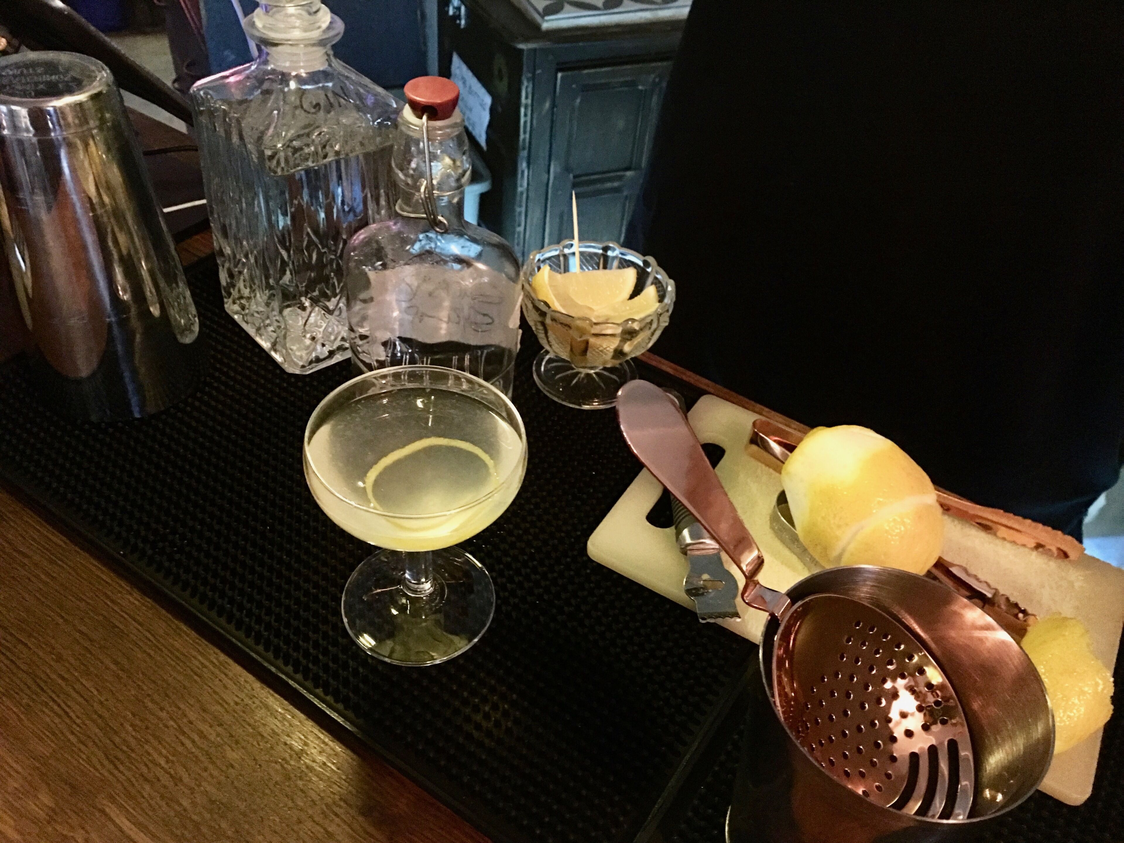 A bar setup with a cocktail glass, a bottle, a bowl of fruit, and a cutting board with a knife and grater.