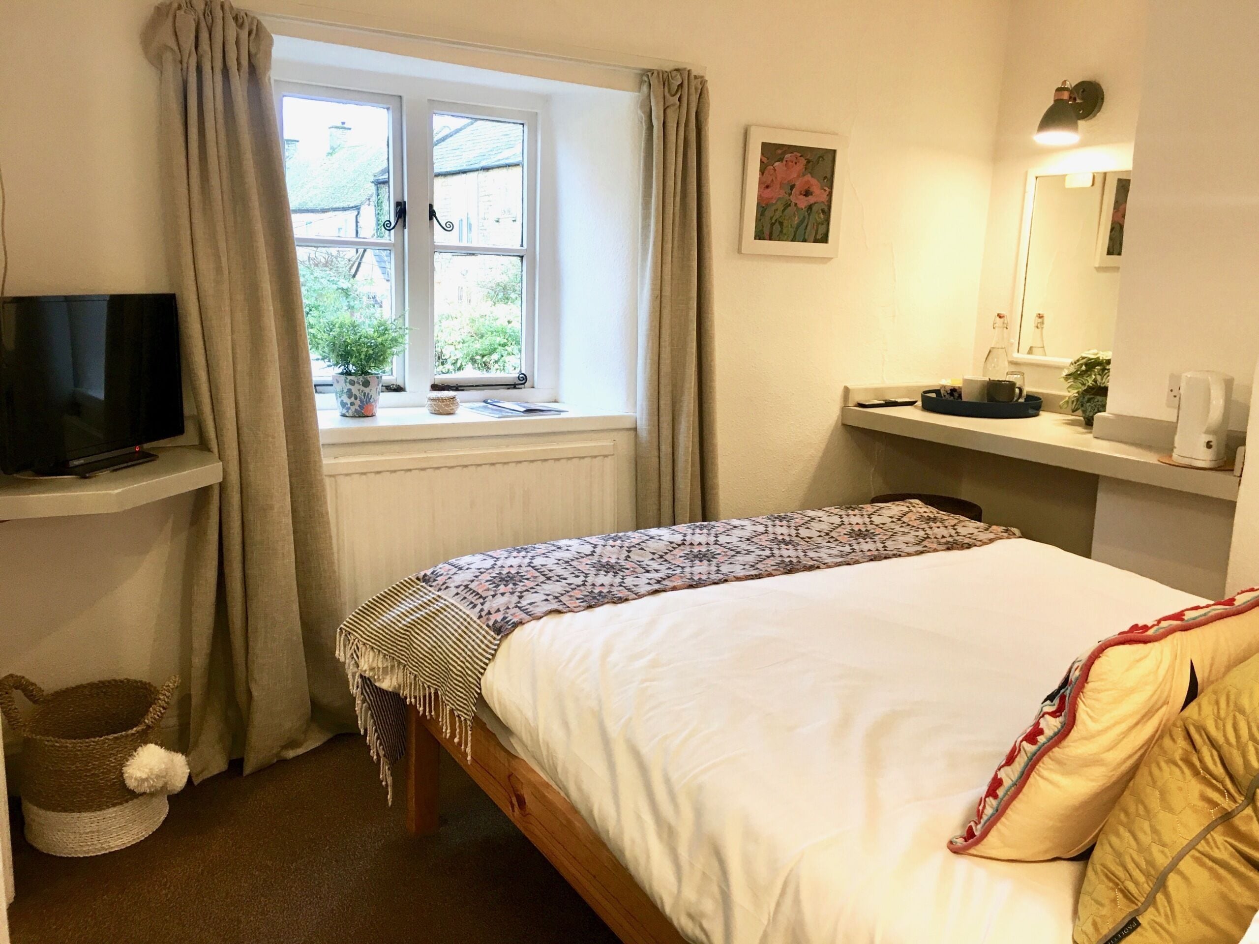 Un dormitorio con una cama, un televisor sobre un mueble bajo, una ventana con cortinas, una mesita de noche con una lámpara y una planta pequeña.