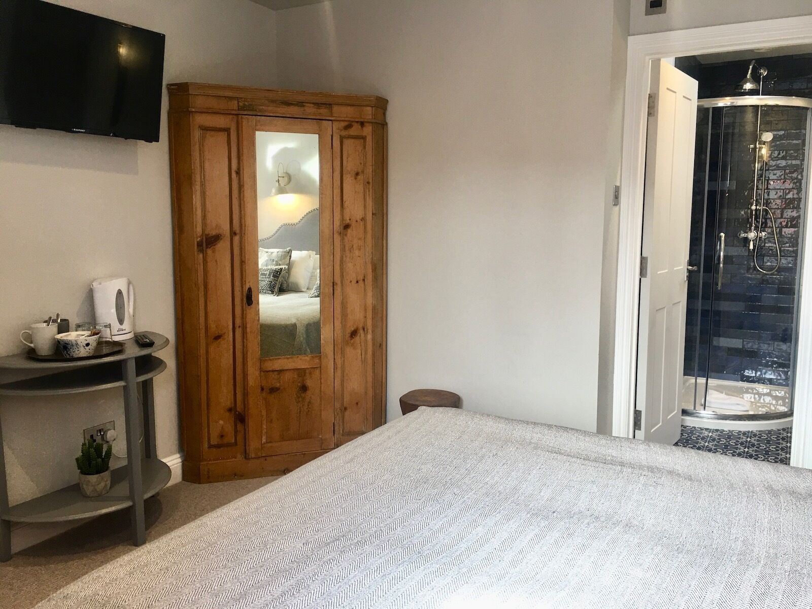 A bedroom with a bed, a wooden wardrobe, a small table with a kettle, and a TV mounted on the wall.