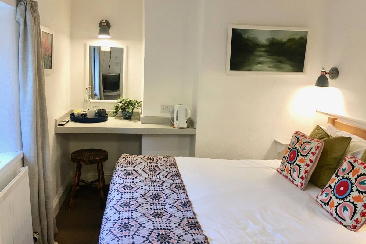 Quarto de hotel com uma cama, uma escrivaninha com cadeira, um espelho, um abajur e um quadro na parede.