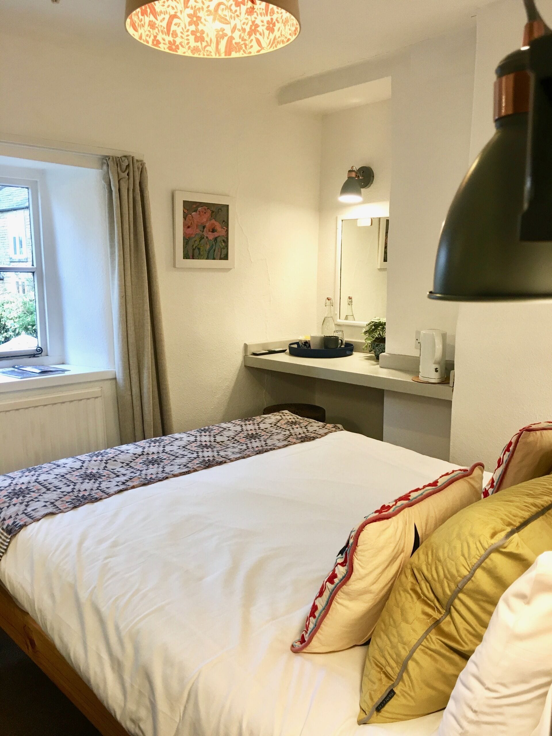 A neatly made bed with patterned bedding, a bedside table with a lamp, a window with curtains, and a painting on the wall.