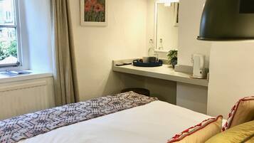 Cama bem arrumada com roupa de cama estampada, mesa de cabeceira com abajur, janela com cortinas e um quadro na parede.