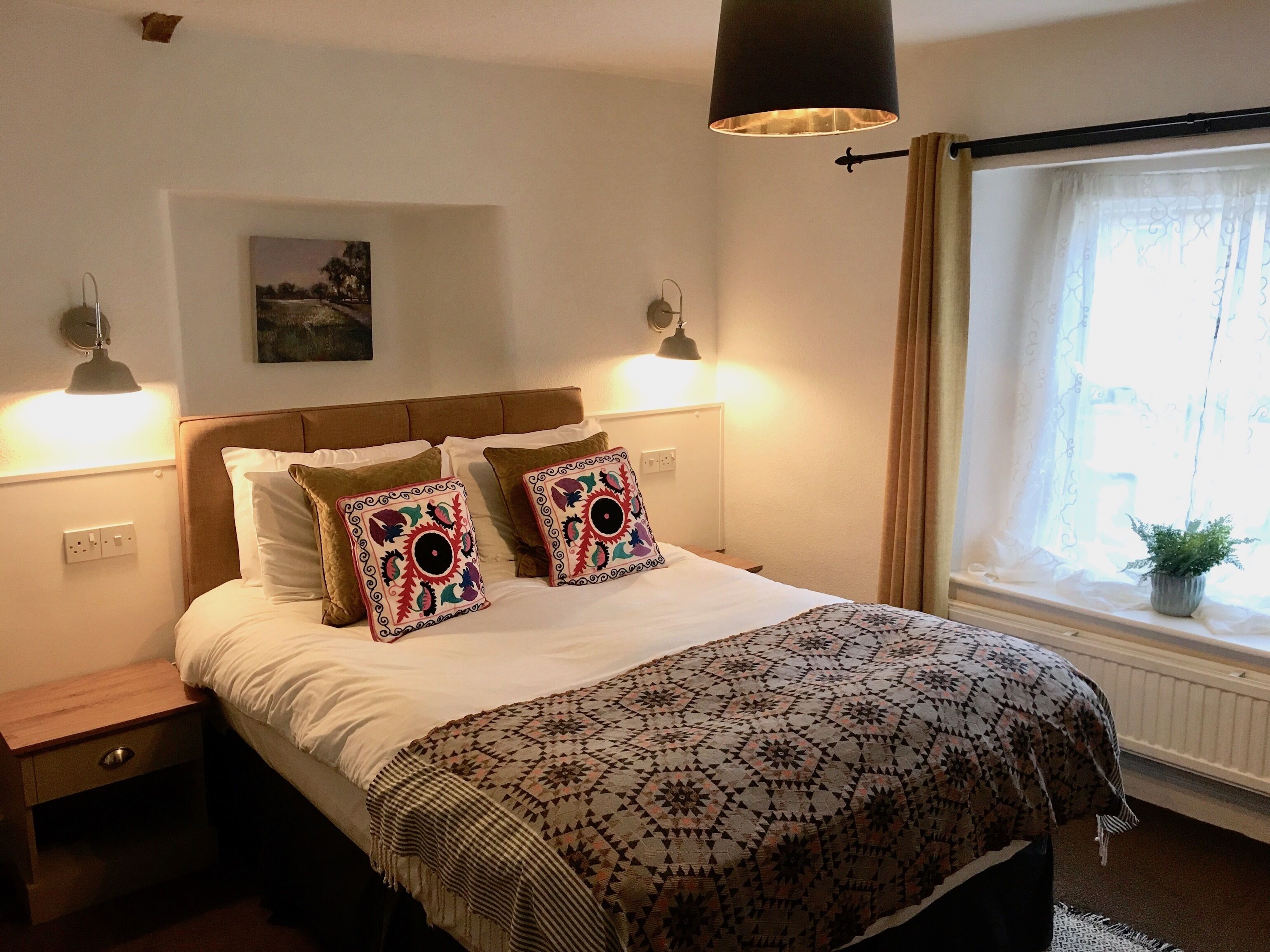 A bedroom with a bed, two decorative pillows, a patterned blanket, bedside lamps, a wall-mounted picture, and a window with curtains.