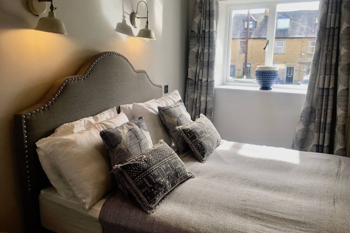 Uma cama bem arrumada com cabeceira, várias almofadas e uma janela com cortinas.