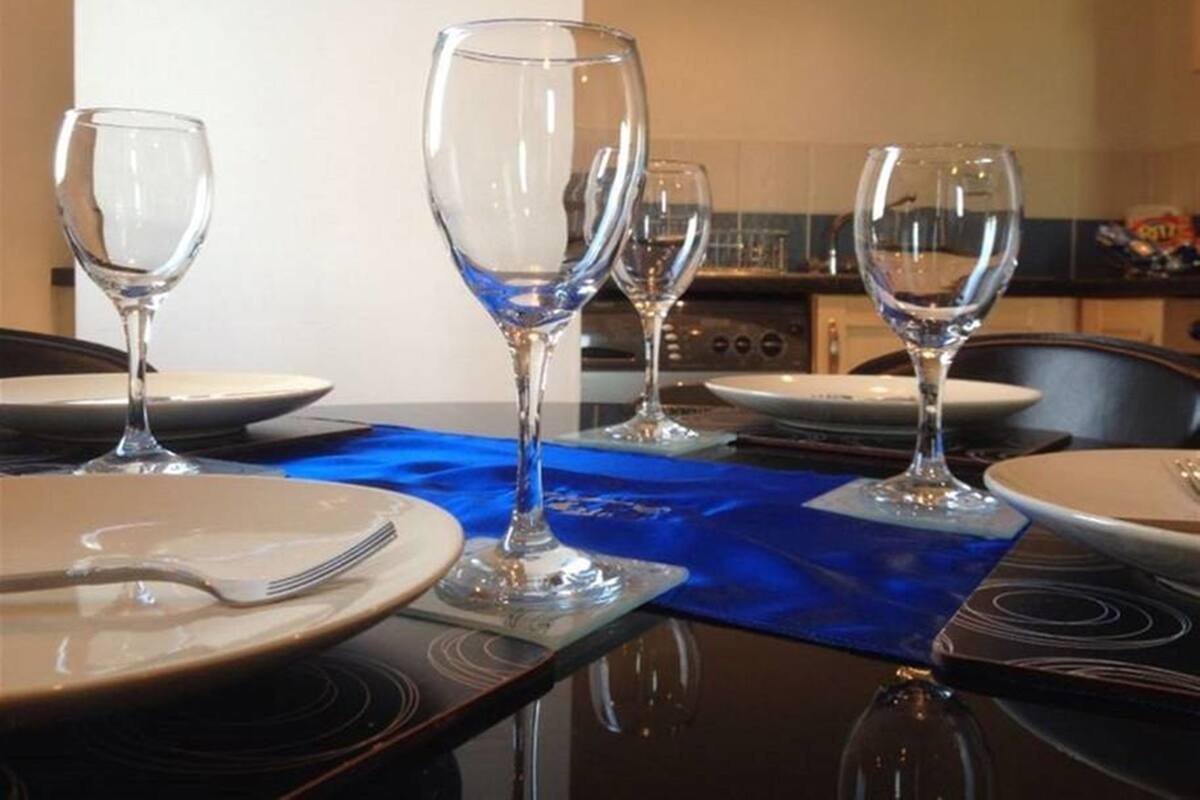A dining table set with four wine glasses and plates, a blue placemat, and a kitchen in the background.