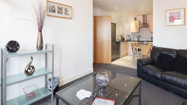 A modern living room with a glass coffee table, a black leather sofa, and a kitchen in the background. - My-Places Serviced Apartments (Manchester)
