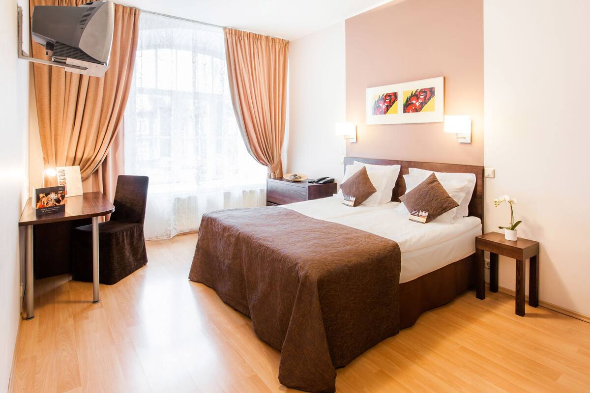 Ett hotellrum med en stor säng, ett skrivbord, en stol, en tv och ett fönster med gardiner.