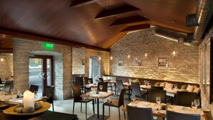 Restaurante con paredes de piedra, vigas de madera e iluminación moderna. Mesas dispuestas con copas de vino y platos.