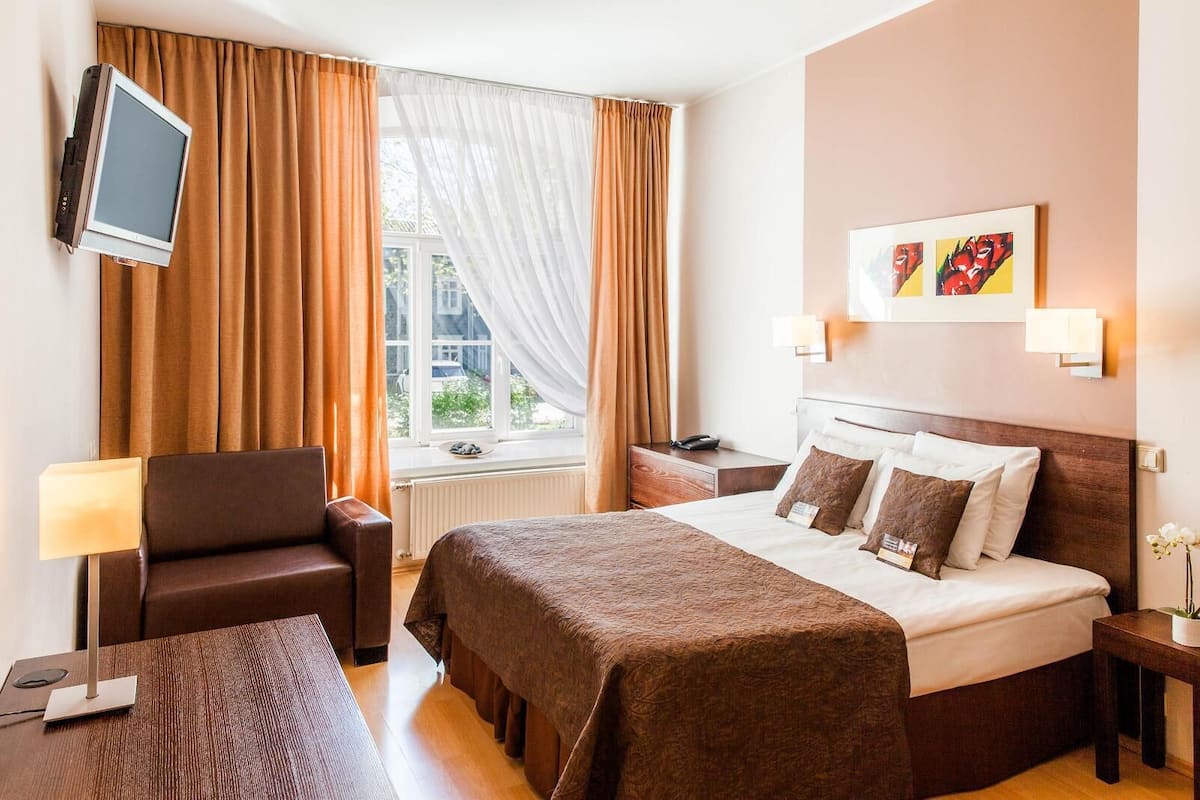 Une chambre d’hôtel avec un lit, une chaise, un bureau, une télévision et une fenêtre avec des rideaux.