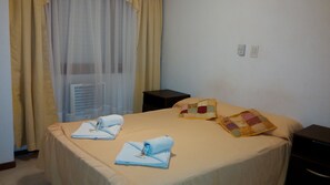 A neatly made bed with a beige bedspread, two folded towels, and a quilted pillow. A small black nightstand is beside the bed. - San Remo Viking Hotel (Pinamar)