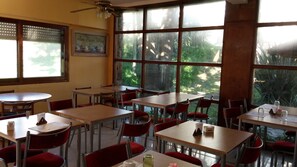 A dining area with tables and chairs, large windows with a view of greenery, and a ceiling fan. - San Remo Viking Hotel (Pinamar)