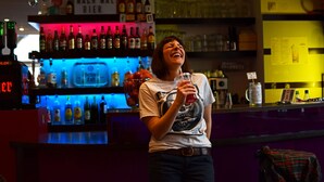 Zona de bar con estantes llenos de botellas y vasos, un cartel que dice 'DIGG MACH MAL BIER', y una persona sosteniendo una bebida.