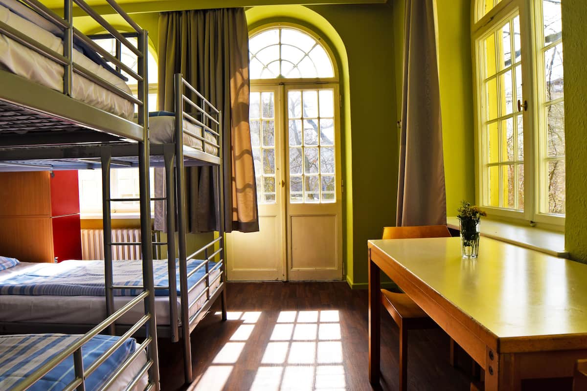A dormitory room with bunk beds, a wooden table, a window with curtains, and a door.
