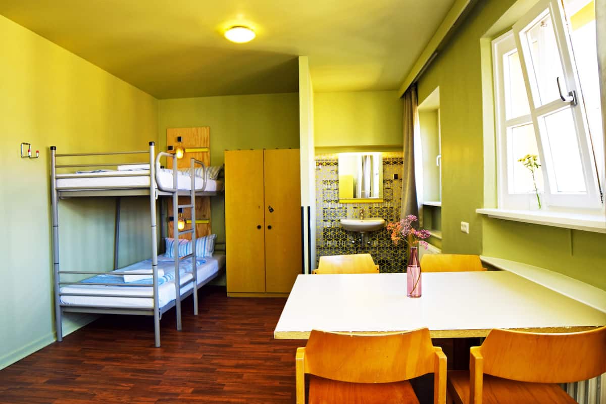 A bunk bed, a wooden wardrobe, a white table with chairs, a sink, and a window with curtains.
