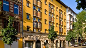 Un edificio amarillo con varias ventanas y balcones, una entrada de piedra y una calle con coches y árboles.