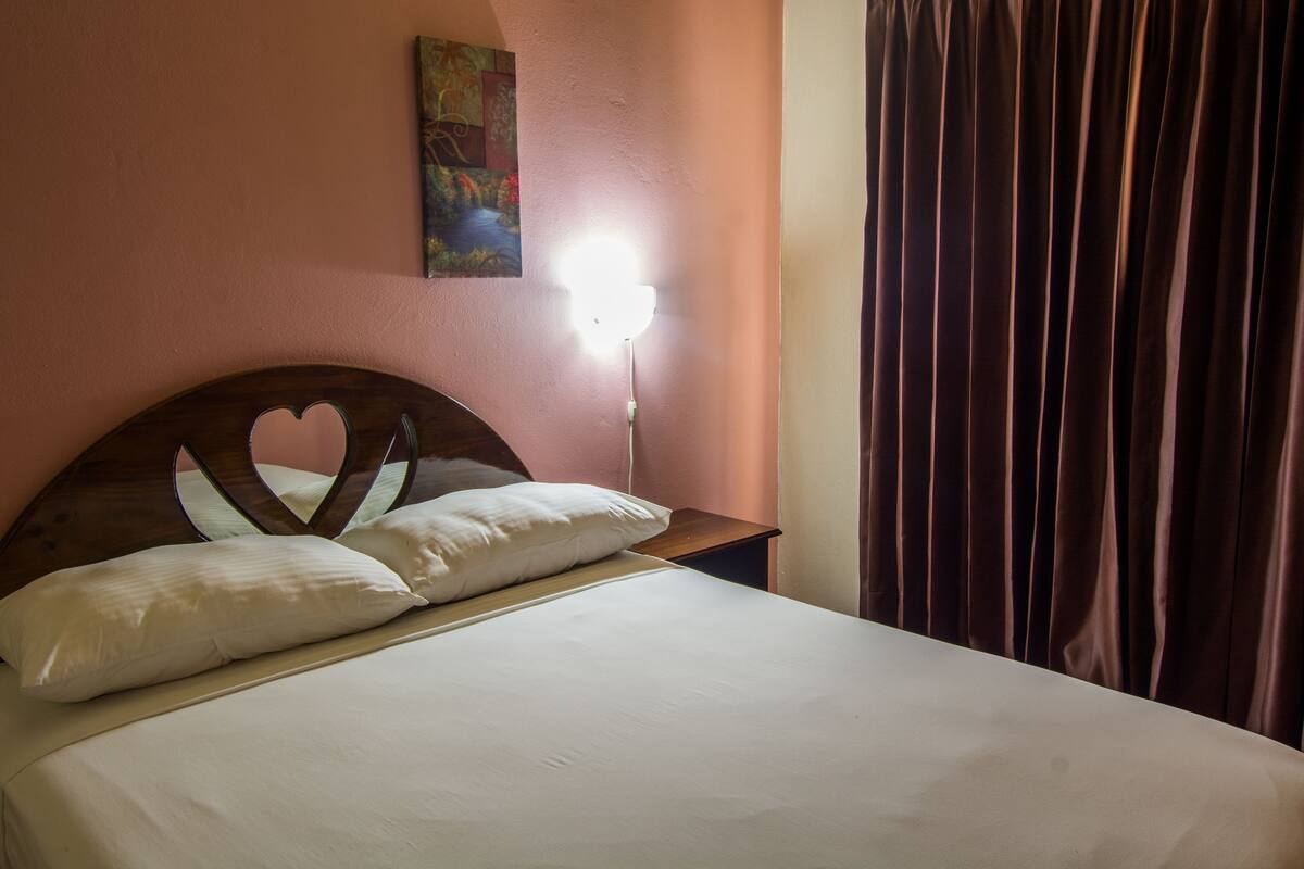 A bed with a heart-shaped headboard, white bedding, a bedside lamp, and a painting on the wall.