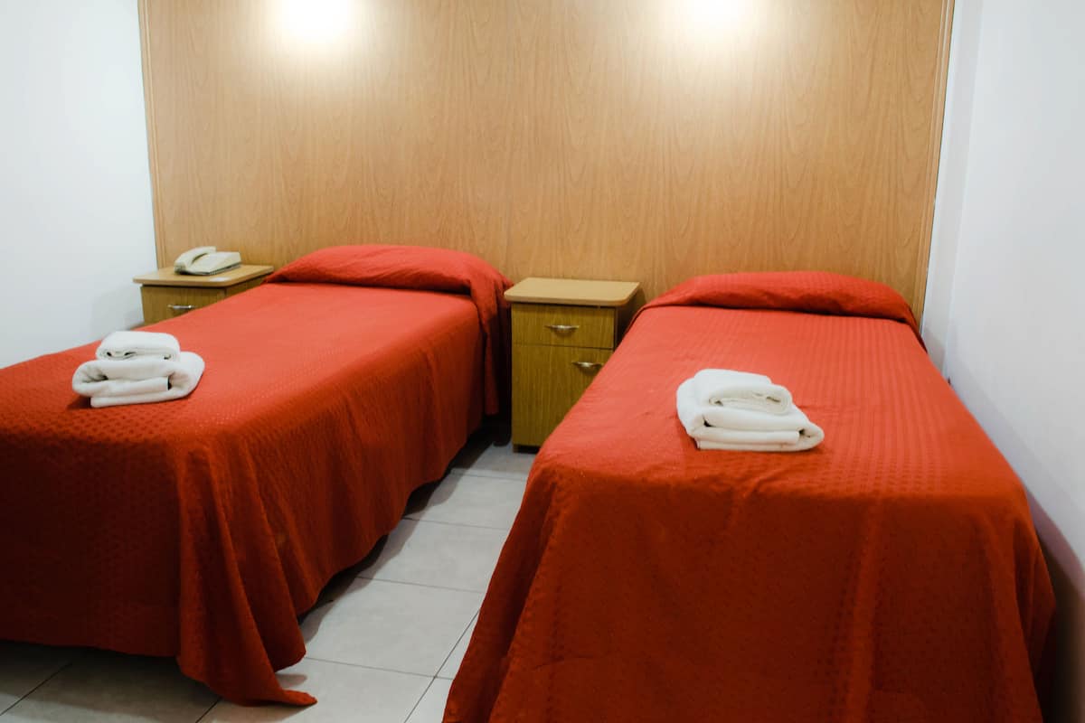 A hotel room with two single beds, each with a red bedspread and a white towel folded on top. There is a wooden nightstand with a telephone between the beds.