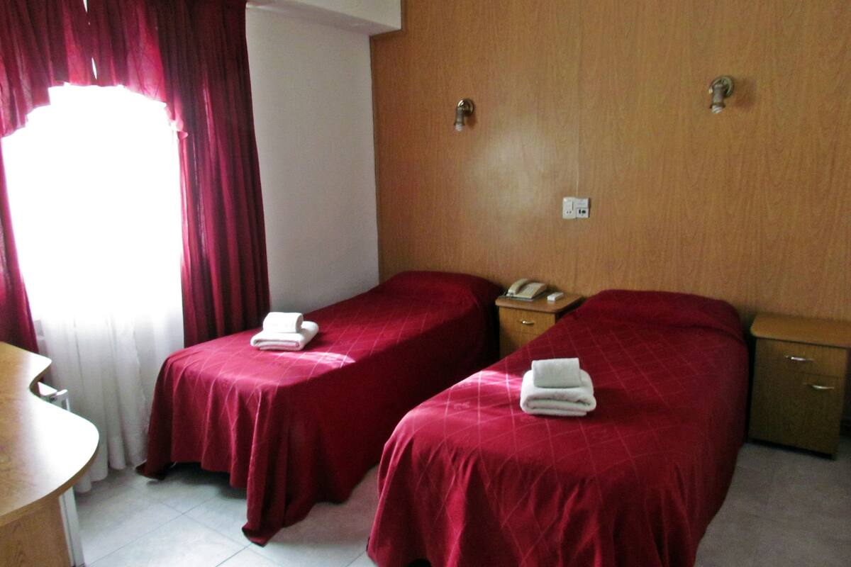 A hotel room with two single beds, a wooden nightstand with a phone, a desk, and a window with red curtains.