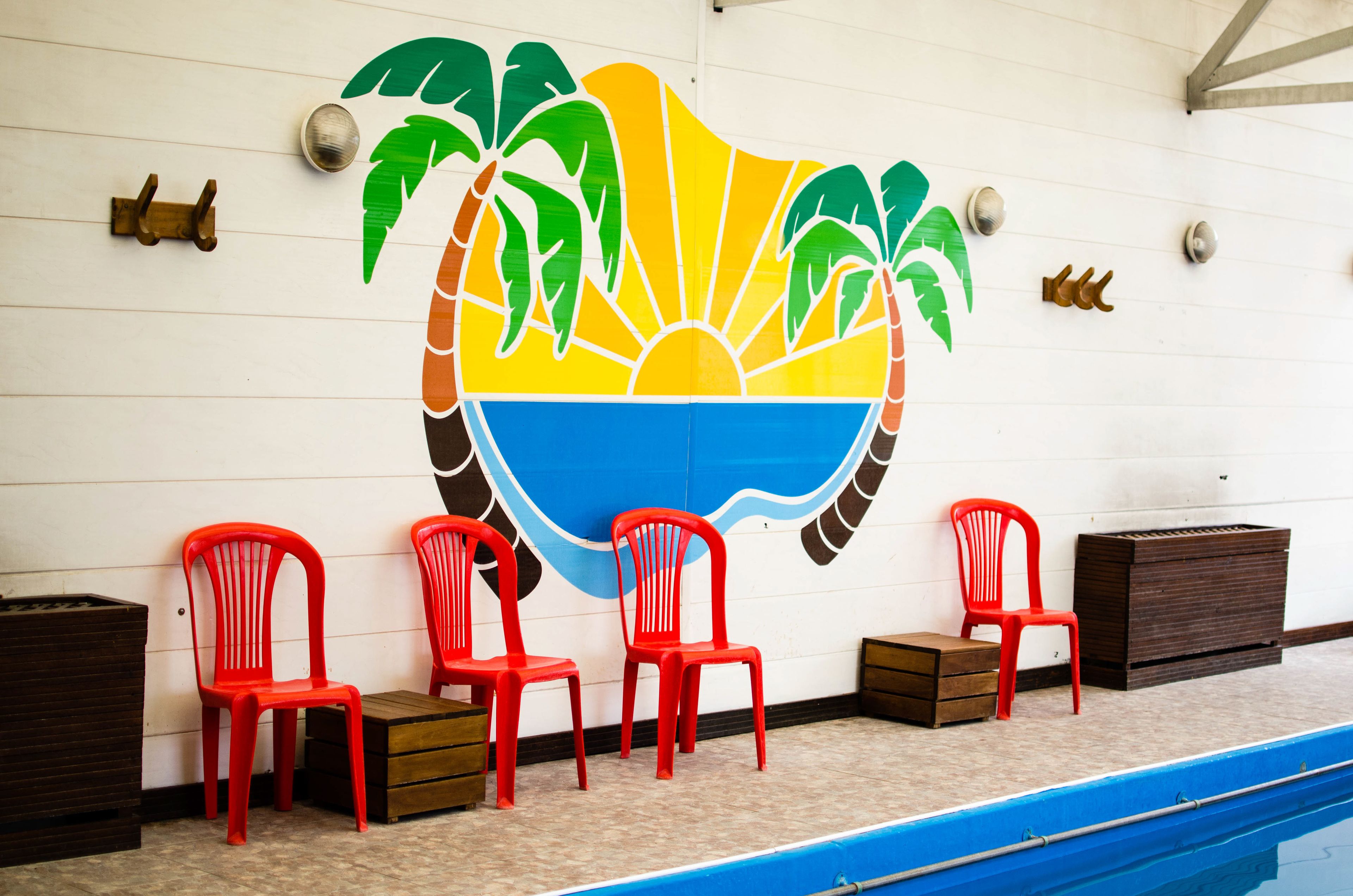 Un mural en la pared con una escena tropical, palmeras y un sol. Hay cuatro sillas de plástico rojas y dos bancos de madera a lo largo de la piscina.