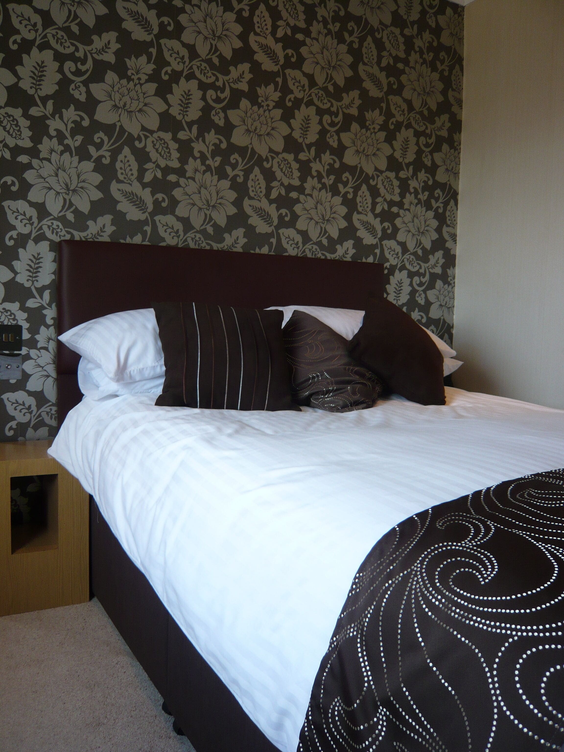 A neatly made bed with white and dark brown bedding, a wooden nightstand, and a floral-patterned wallpaper.