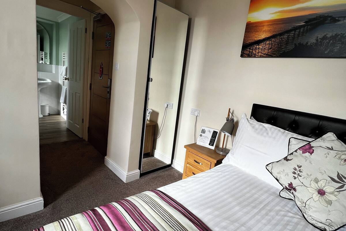 Een hotelkamer met een bed, een schilderij van een zonsondergang boven een pier, en een uitzicht op een badkamer door een boog.