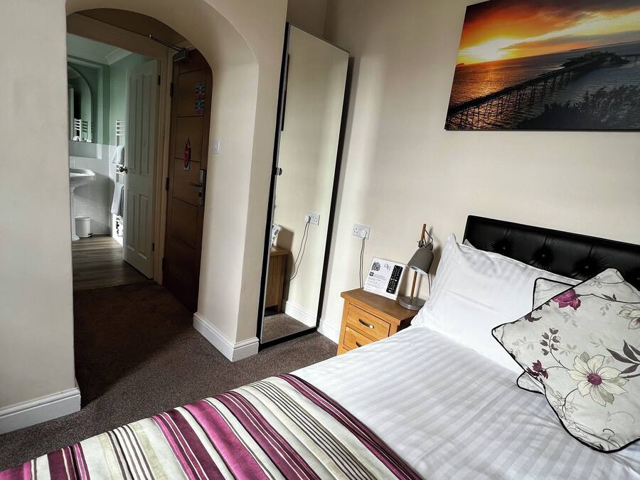 A hotel room with a bed, a painting of a sunset over a pier, and a view of a bathroom through an archway.