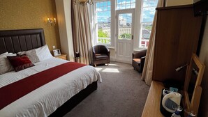 A hotel room with a large bed, a wooden desk, a chair, a television, and a view of the city through the windows. - Queenswood Hotel (Weston-super-Mare)