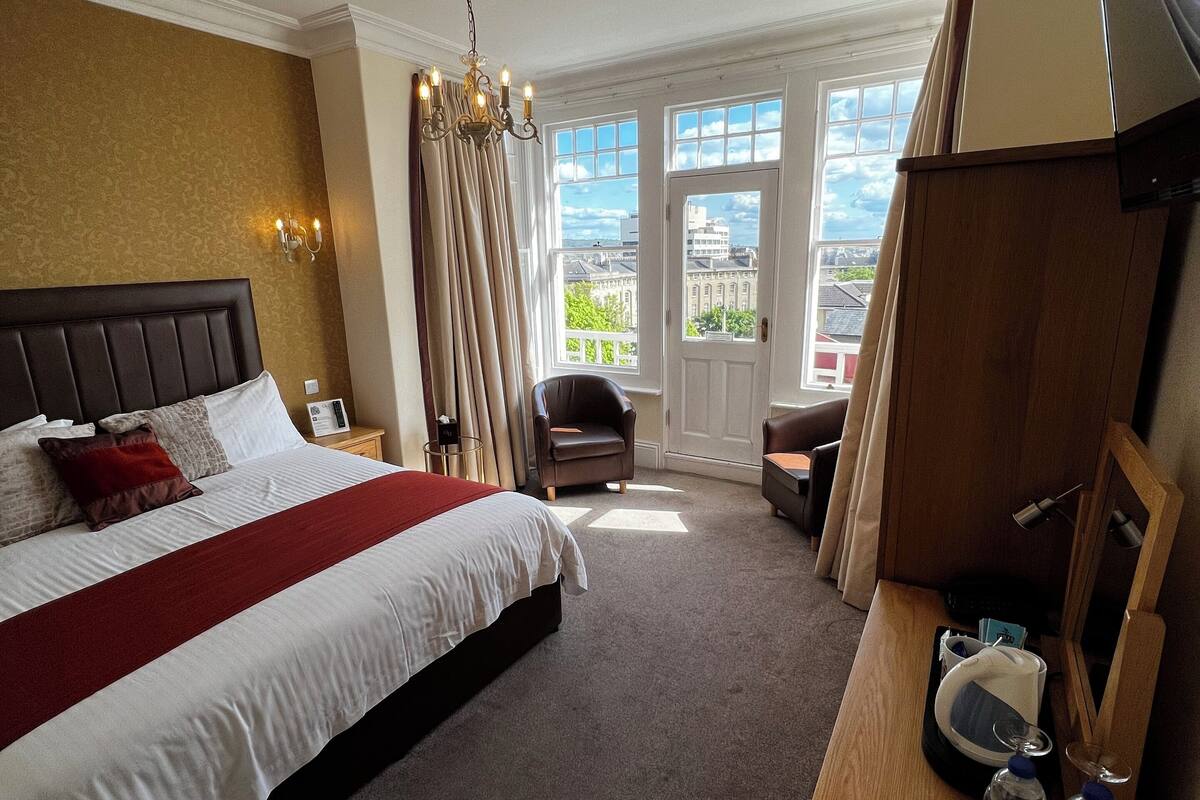 Een hotelkamer met een groot bed, een houten bureau, een stoel, een televisie en uitzicht op de stad door de ramen.