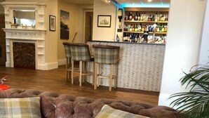 A brown leather sofa with plaid cushions, a fireplace, a bar area with stools, and a plant. - Queenswood Hotel (Weston-super-Mare)
