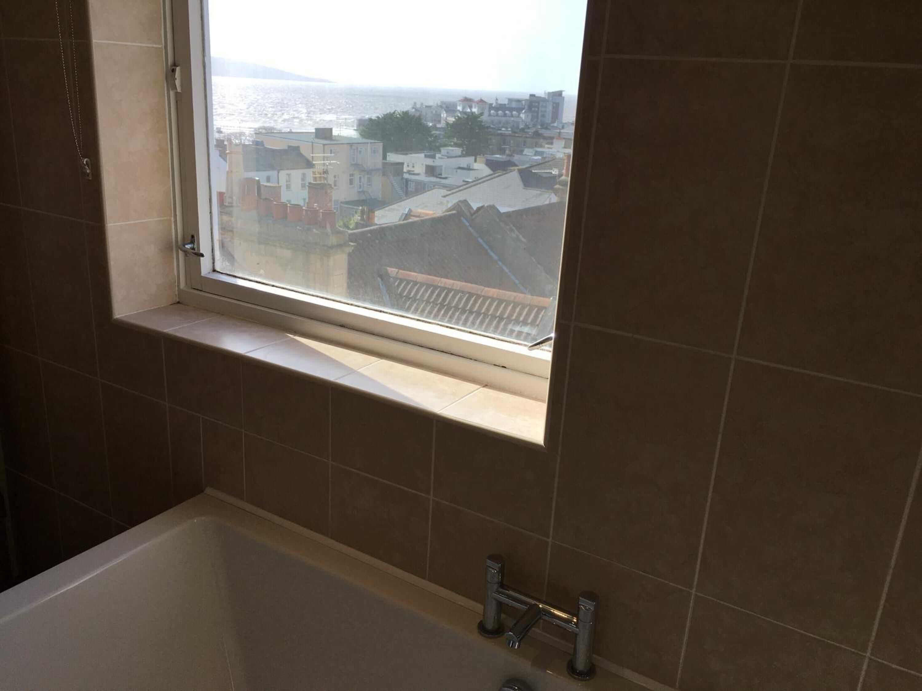 A bathroom with a bathtub, a window with a view of buildings, and a tiled wall.