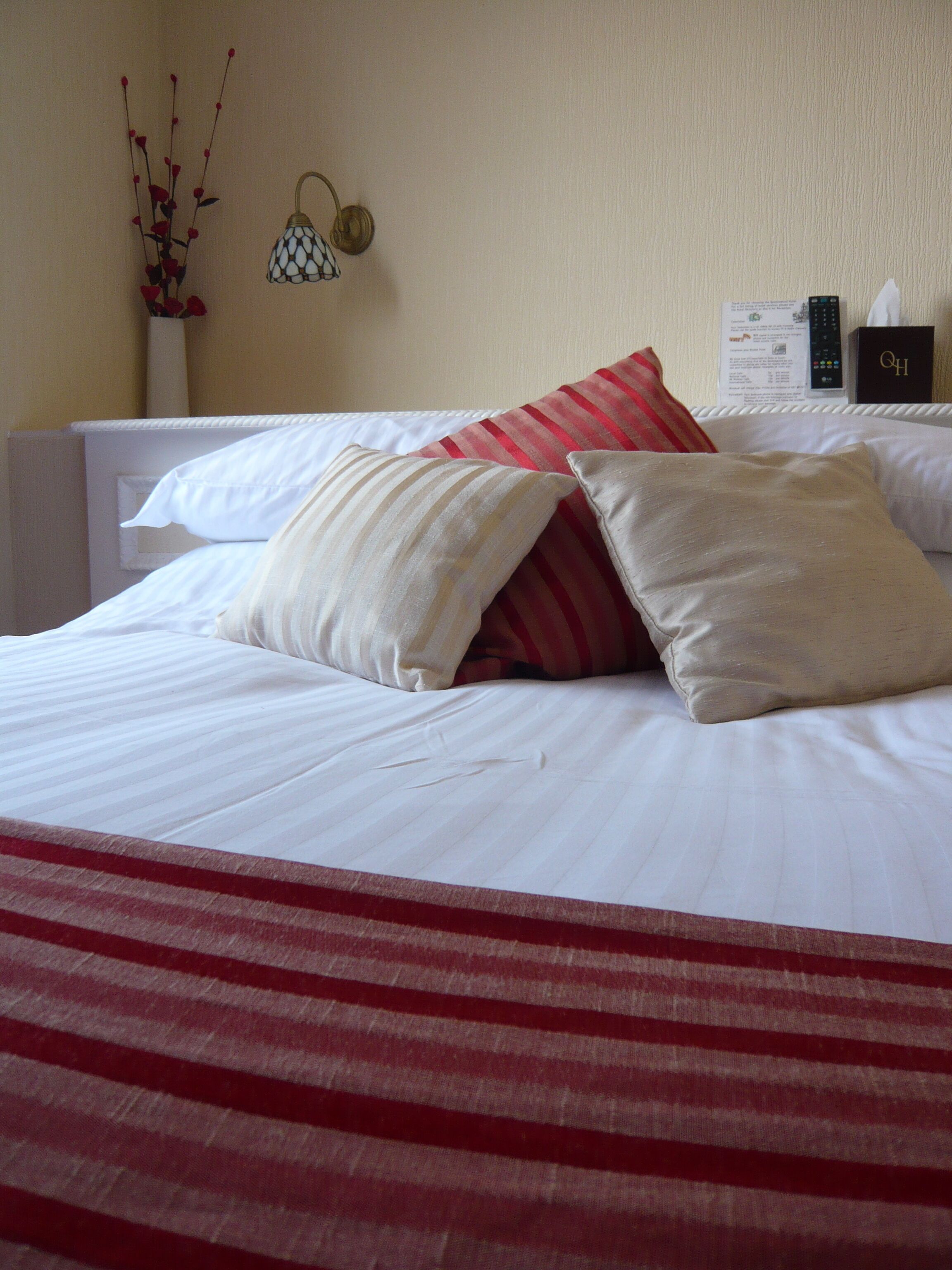 Een net opgemaakt bed met witte lakens en rode gestreepte kussens, een wandlamp en een afstandsbediening op het nachtkastje.