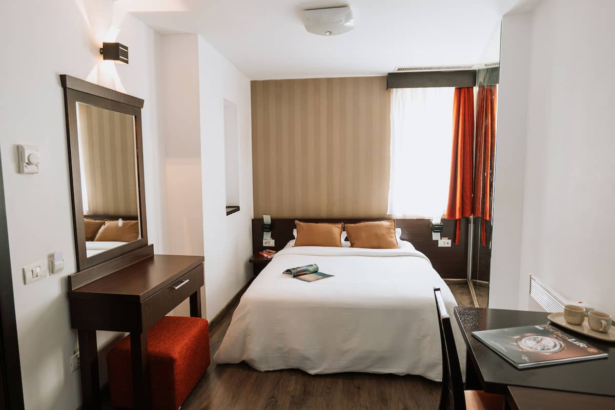 A hotel room with a bed, a desk, a chair, a mirror, a nightstand, and a window with curtains.