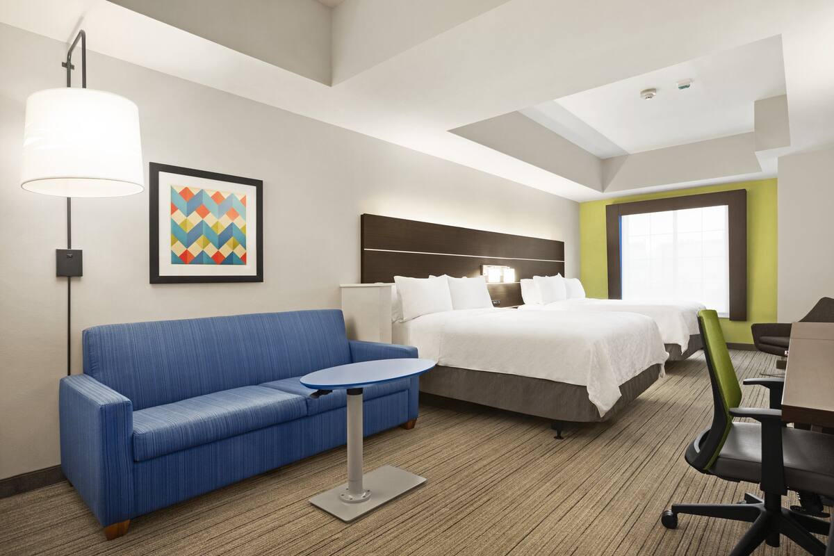 A hotel room with a blue sofa, a small round table, a bed with white bedding, a headboard, and a large window.