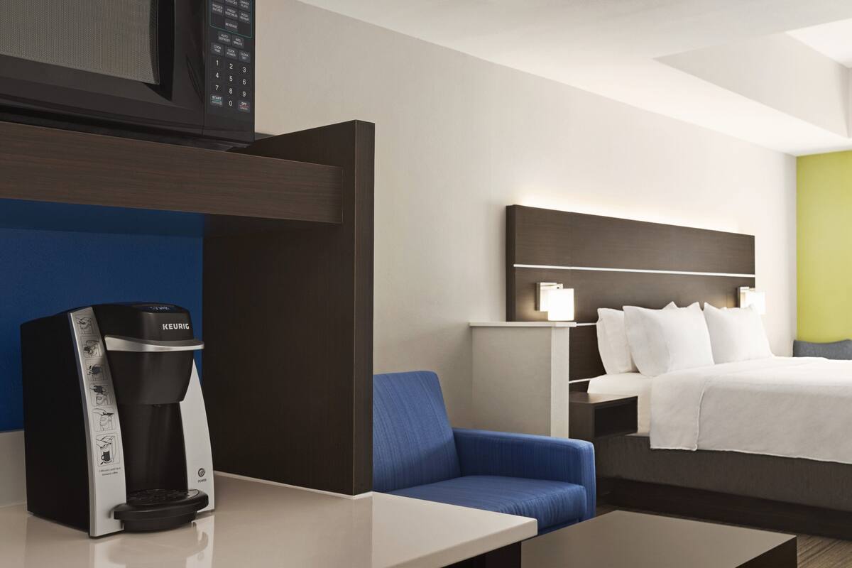 A hotel room with a bed, a blue armchair, a coffee machine, a bedside table, and a nightstand.