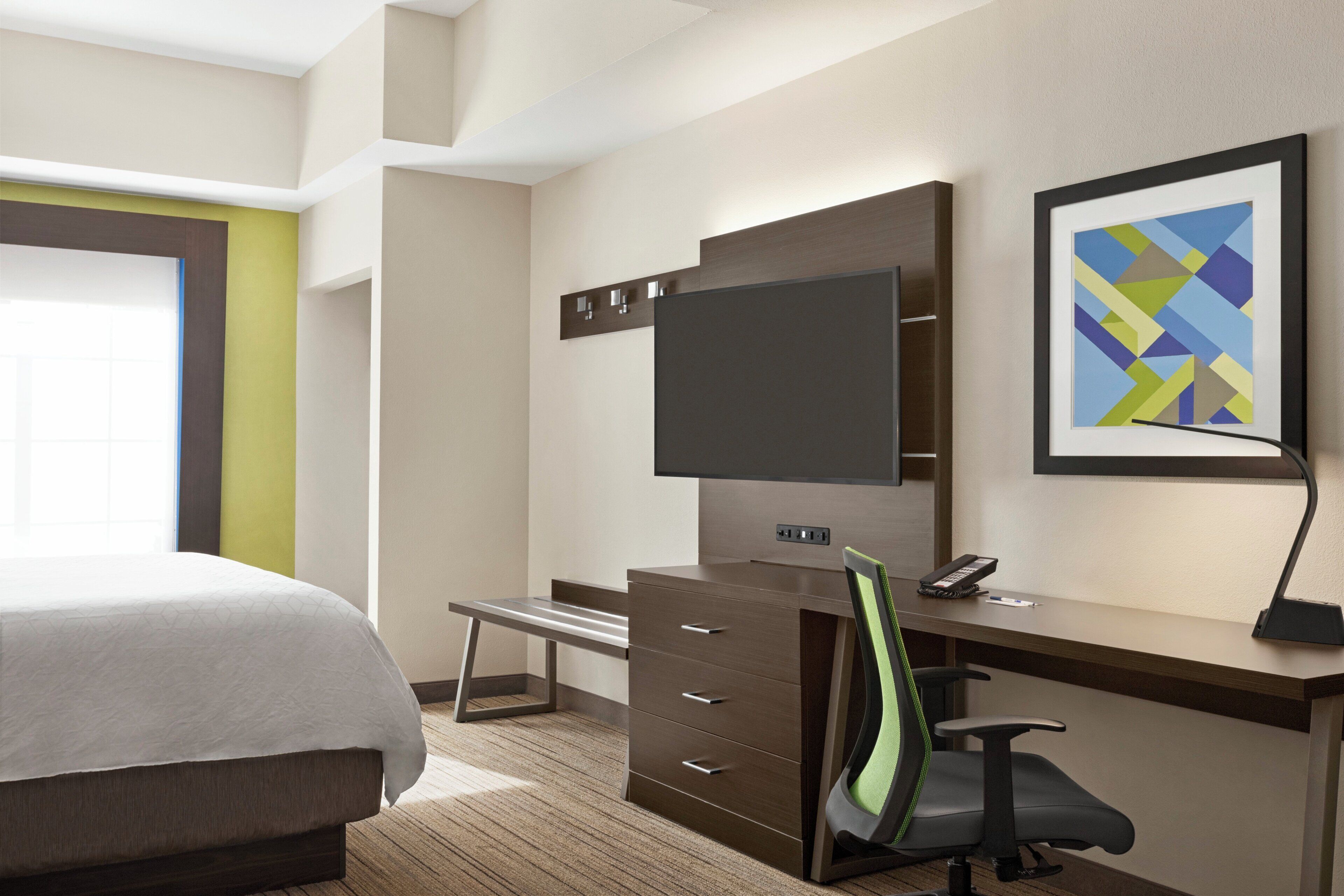 A hotel room with a bed, a desk with a chair, a flat-screen TV, a telephone, and a framed abstract painting on the wall.