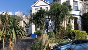 A two-story house with a white facade and blue trim, surrounded by lush greenery and a parked car. - The Brandize Guest House (Torquay)