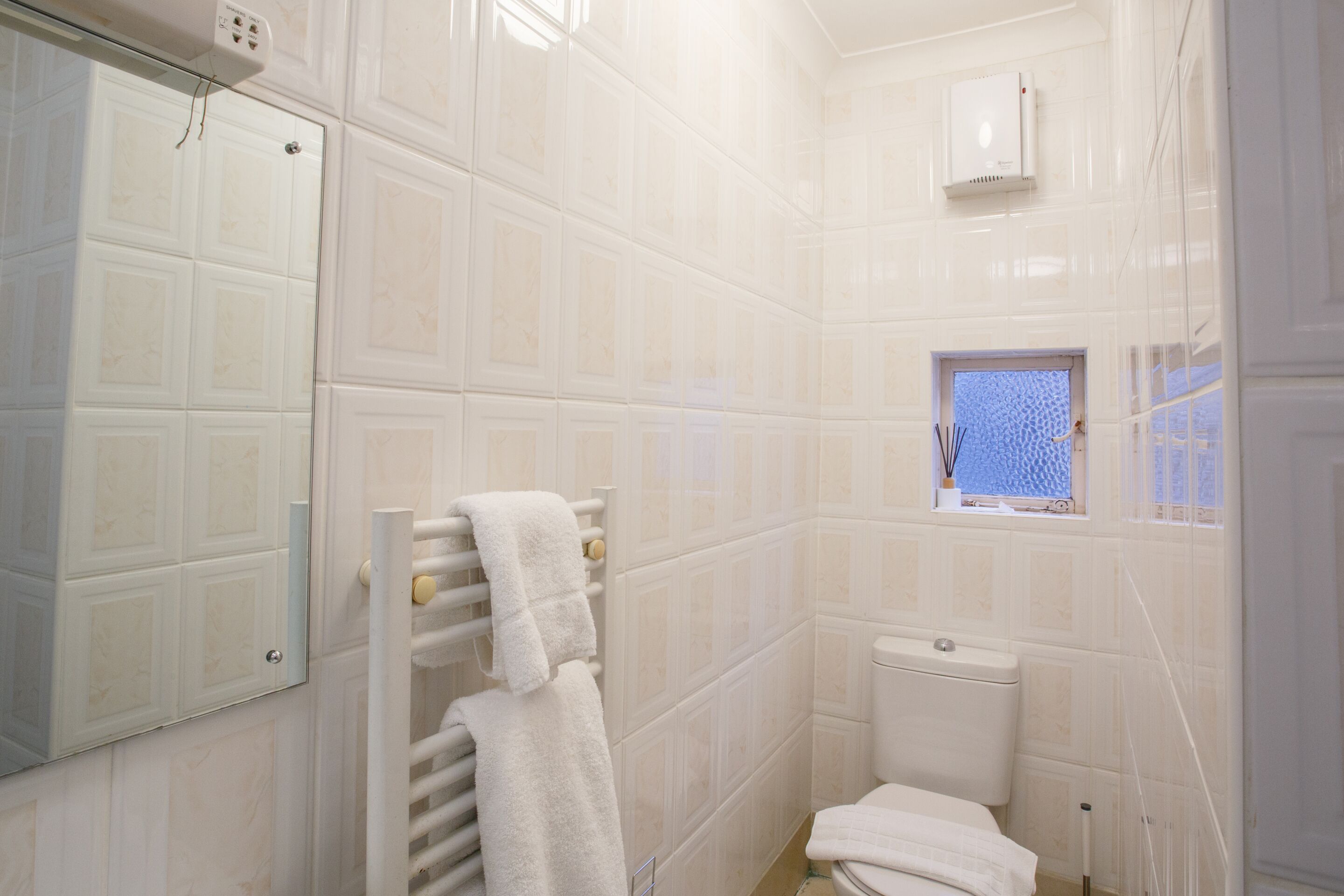 Une salle de bain avec des murs carrelés de carreaux blancs, des toilettes, une fenêtre donnant sur un beau paysage et un porte-serviettes avec une serviette blanche.