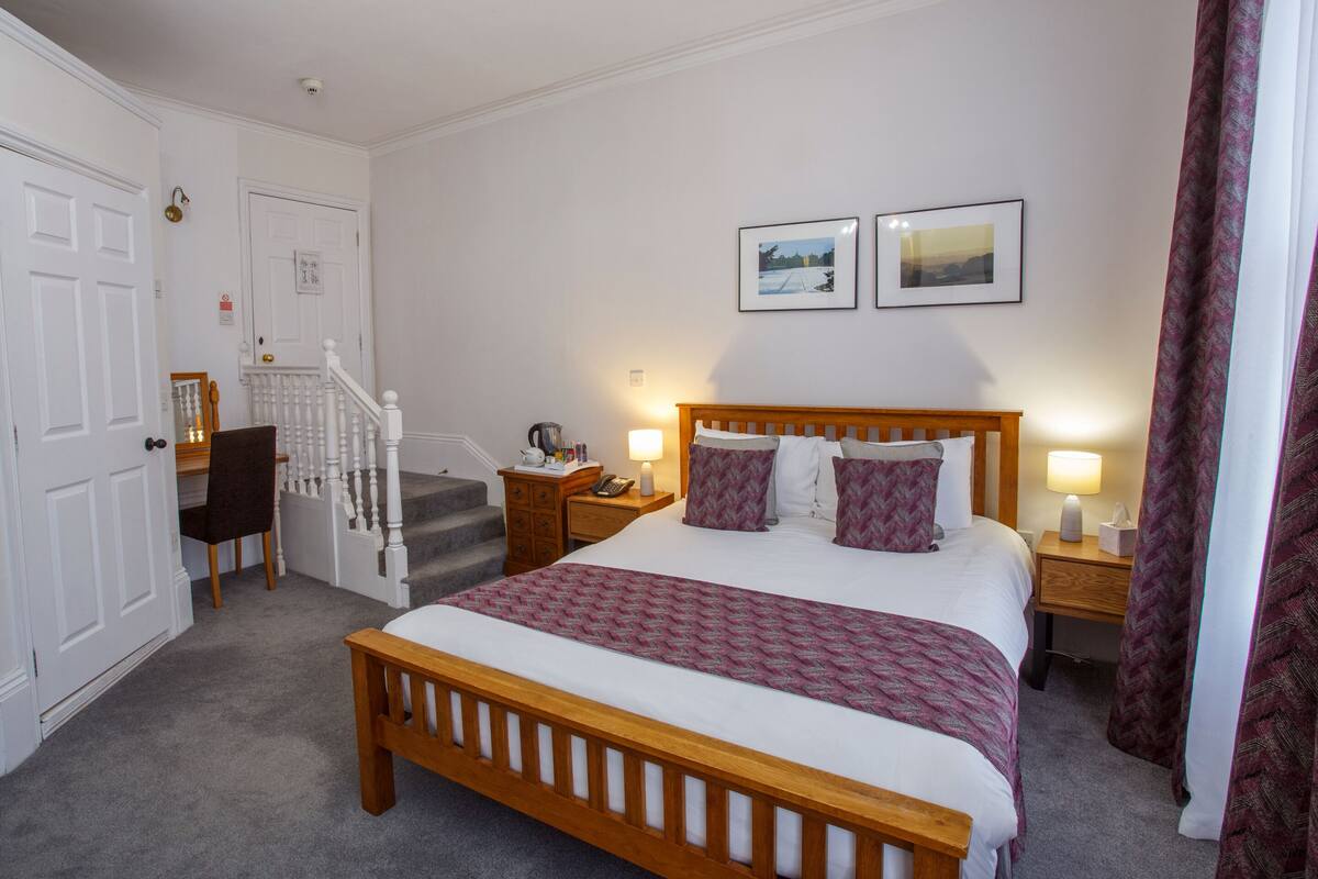 A neatly arranged bedroom with a wooden bed, two bedside tables with lamps, a desk with a chair, a staircase, and two framed pictures on the wall.