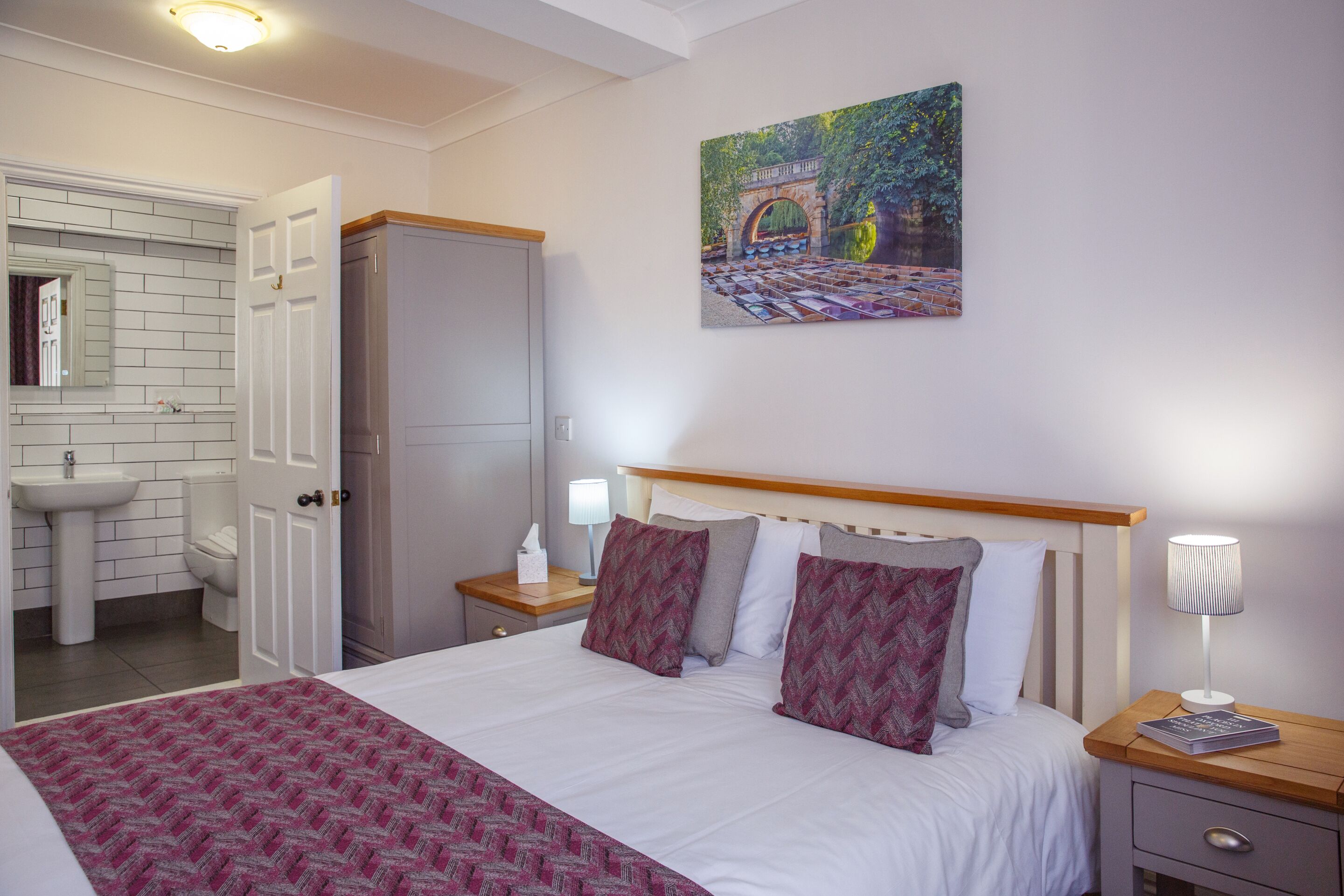 A hotel room with a bed, a nightstand, a lamp, a painting, a bathroom with a sink and toilet, and a wardrobe.